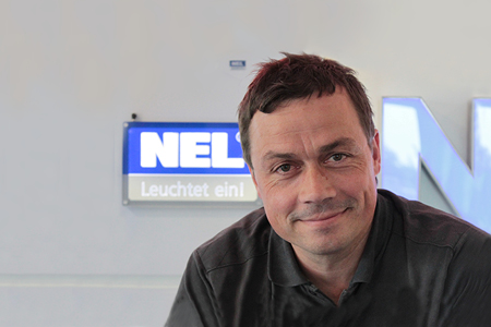 Achim Hammer, Mitarbeiter bei NEL, in einem dunkelgrauen Hemd, steht vor einer NEL-Leuchttafel und lächelt in die Kamera.