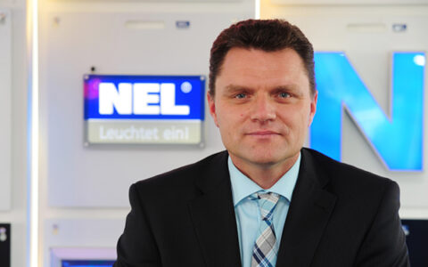 Andreas Kattwinkel im Anzug sitzt vor einer beleuchteten NEL-Werbetafel. Er trägt einen schwarzen Anzug, ein hellblaues Hemd und eine gestreifte Krawatte.