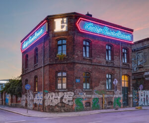 Ein altes Backsteingebäude mit Graffiti an den Wänden, das von neonfarbenen Schriftzügen beleuchtet wird. Der Schriftzug "Leipziger Baumwollspinnerei" von NEL ist in einer roten und blauen Neonleuchtreklame rund um das Dach zu sehen. Vor dem Gebäude sind Straßen und Schilder, während der Himmel im Hintergrund im Abendlicht glüht.