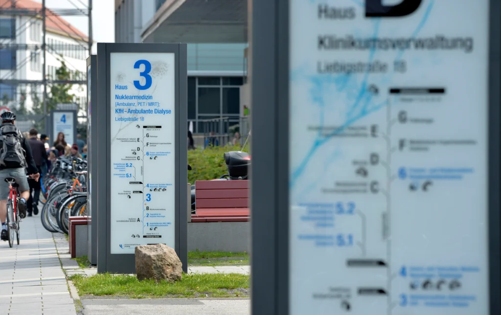 Wegweiser des Leit- und Orientierungssystems am Universitätsklinikum Leipzig, entwickelt und aufgestellt von der NEL. Im Vordergrund ein Schild mit der Aufschrift "Haus 3 - Nuklearmedizin" und weiteren Abteilungen. Im Hintergrund sind Fahrräder und Personen zu sehen, die auf dem Gelände unterwegs sind.