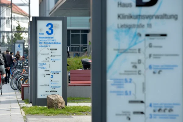 Wegweiser des Leit- und Orientierungssystems am Universitätsklinikum Leipzig, entwickelt und aufgestellt von der NEL. Im Vordergrund ein Schild mit der Aufschrift "Haus 3 - Nuklearmedizin" und weiteren Abteilungen. Im Hintergrund sind Fahrräder und Personen zu sehen, die auf dem Gelände unterwegs sind.