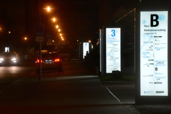 Beleuchtete Wegweiser und Schilder des Leitsytem und Orientierungsskonzepts am Universitätsklinikum Leipzig in der Nacht, entwickelt und aufgestellt von der NEL. Im Vordergrund ist eine beleuchtete Infotafel mit der Aufschrift "Haus B - Klinikumsverwaltung" zu sehen, weitere beleuchtete Schilder mit Wegweisern sind im Hintergrund erkennbar. Auf der Straße fährt ein Taxi, während die Straßenlaternen die Umgebung schwach erleuchten.