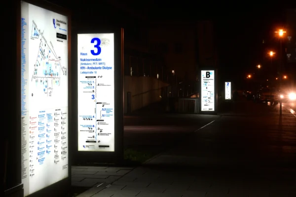 Beleuchtete Wegweiser des Leit- und Orientierungssystems am Universitätsklinikum Leipzig in der Nacht. Das Schild für "Haus 3" weist auf die Abteilungen Nuklearmedizin und ambulante Dialyse hin. Weitere Schilder sind entlang des Gehwegs zu sehen, und im Hintergrund fahren Autos mit eingeschaltetem Licht.