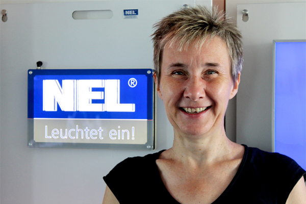 Kerstin Schumann steht vor einem beleuchteten NEL-Logo, lächelnd und in schwarzem Oberteil.