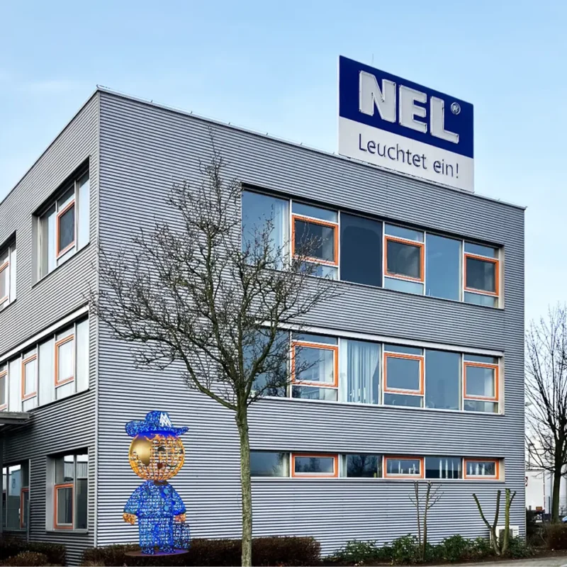 Außenansicht eines modernen Bürogebäudes der Firma NEL mit silberner Metallfassade und rechteckigen Fenstern, die orange umrahmt sind. Auf dem Dach befindet sich ein großes NEL-Logo mit dem Slogan „Leuchtet ein!“.