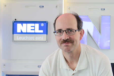 Michael Herrmann, Mitarbeiter von NEL, in einem weißen Poloshirt, steht vor einer NEL-Leuchttafel und lächelt in die Kamera.