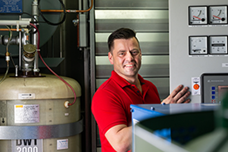 Nico Partschefeld, Mitarbeiter bei NEL, in einem roten Poloshirt, steht lächelnd vor technischen Anlagen und Bedienelementen.