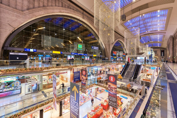 Innenansicht des Leipziger Hauptbahnhofs mit festlicher Weihnachtsdekoration. Mehrere Etagen voller Geschäfte und Stände sind zu sehen, während die oberen Ebenen des Bahnhofsbereichs mit glitzernden Lichterketten und goldenen Sternen geschmückt sind. Menschen gehen durch die Einkaufspassage, und an den Gleisen warten Züge.