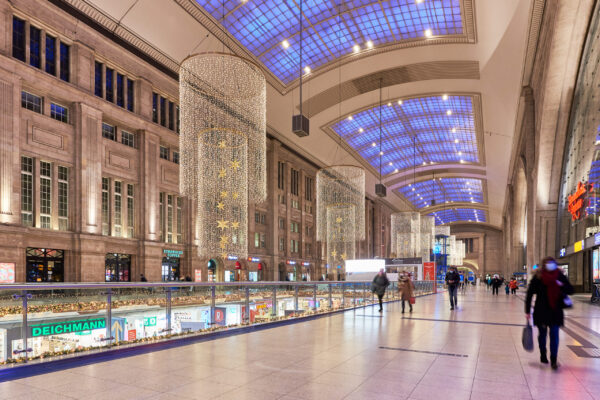 Weitläufige Halle des Leipziger Hauptbahnhofs, geschmückt mit großen, zylindrischen Lichterketten, die von der hohen Decke hängen. Entlang der Balustraden sind ebenfalls Weihnachtsdekorationen zu sehen. Unten sind Geschäfte, während Passanten durch die obere Etage gehen.