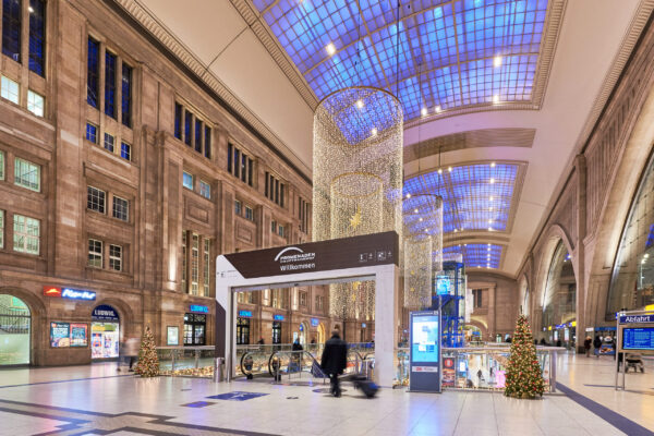 Innenansicht des Leipziger Hauptbahnhofs mit festlicher Weihnachtsdekoration. Glitzernde Lichterketten und Weihnachtsbäume schmücken die große Halle mit ihrer hohen Decke und den imposanten Glasfenstern. Im Vordergrund befindet sich ein „Willkommen“-Schild der Promenaden, im Hintergrund sind Geschäfte und Rolltreppen zu sehen.