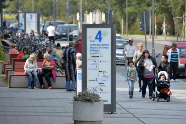Menschen laufen und sitzen auf einer gepflasterten Straße vor dem Universitätsklinikum Leipzig. Im Vordergrund steht eine große Informationssäule mit einem Lageplan für das Klinikgebäude 4. Im Hintergrund sind Fahrräder, Bänke und weitere Passanten zu sehen.