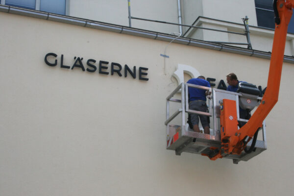 Außenansicht eines Gebäudes mit dem Schriftzug „GLÄSERNE 3D FABRIK by Rapidobject“. Der 3D-Schriftzug mit einer rot-braunen, wabenartigen Struktur und weißen Elementen wird von NEL montiert.