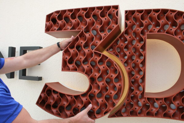 Zwei Hände montieren einen großen 3D-gedruckten Buchstaben "3" mit einer wabenartigen Struktur an einer Wand. Der Buchstabe ist Teil eines Schildes und wird präzise angebracht.