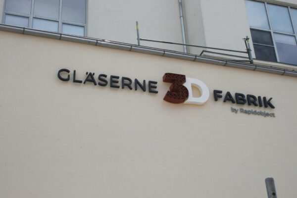 Außenansicht eines Gebäudes mit dem Schriftzug „GLÄSERNE 3D FABRIK by Rapidobject“. Der 3D-Schriftzug mit einer rot-braunen, wabenartigen Struktur und weißen Elementen wird von NEL montiert.