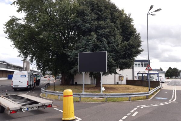 Eine leere, schwarze Anzeigetafel steht neben einem großen Baum auf einem Firmengelände. Im Hintergrund sind mehrere Fahrzeuge, darunter ein Transporter von NEL, sowie industrielle Gebäude zu sehen. Ein Anhänger steht auf der Straße, die von Fahrbahnmarkierungen und Verkehrszeichen gesäumt ist.