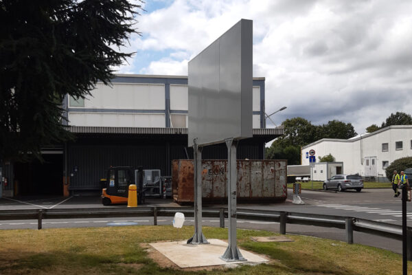 Rückansicht einer neu installierten Anzeigetafel auf einem Firmengelände. Die Tafel steht auf einem Betonfundament, und im Hintergrund sind industrielle Gebäude, ein Gabelstapler und ein großer Container zu sehen. Der Himmel ist teils bewölkt, teils blau.