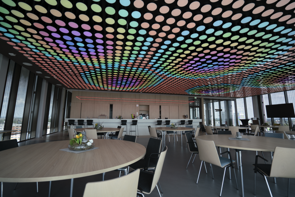 Ein moderner Raum mit einer auffälligen Lichtdecke, die aus zahlreichen farbigen, kreisförmigen Lichtern besteht. Die Decke leuchtet in verschiedenen Farbtönen, darunter Blau, Grün, Rosa und Gelb. Der Raum ist mit runden Holztischen und schwarzen sowie hellen Stühlen ausgestattet. Im Hintergrund befindet sich eine Theke, und große Fenster lassen viel Tageslicht herein.