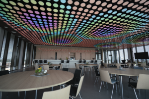 Ein moderner Raum mit einer auffälligen Lichtdecke, die aus zahlreichen farbigen, kreisförmigen Lichtern besteht. Die Decke leuchtet in verschiedenen Farbtönen, darunter Blau, Grün, Rosa und Gelb. Der Raum ist mit runden Holztischen und schwarzen sowie hellen Stühlen ausgestattet. Im Hintergrund befindet sich eine Theke, und große Fenster lassen viel Tageslicht herein.