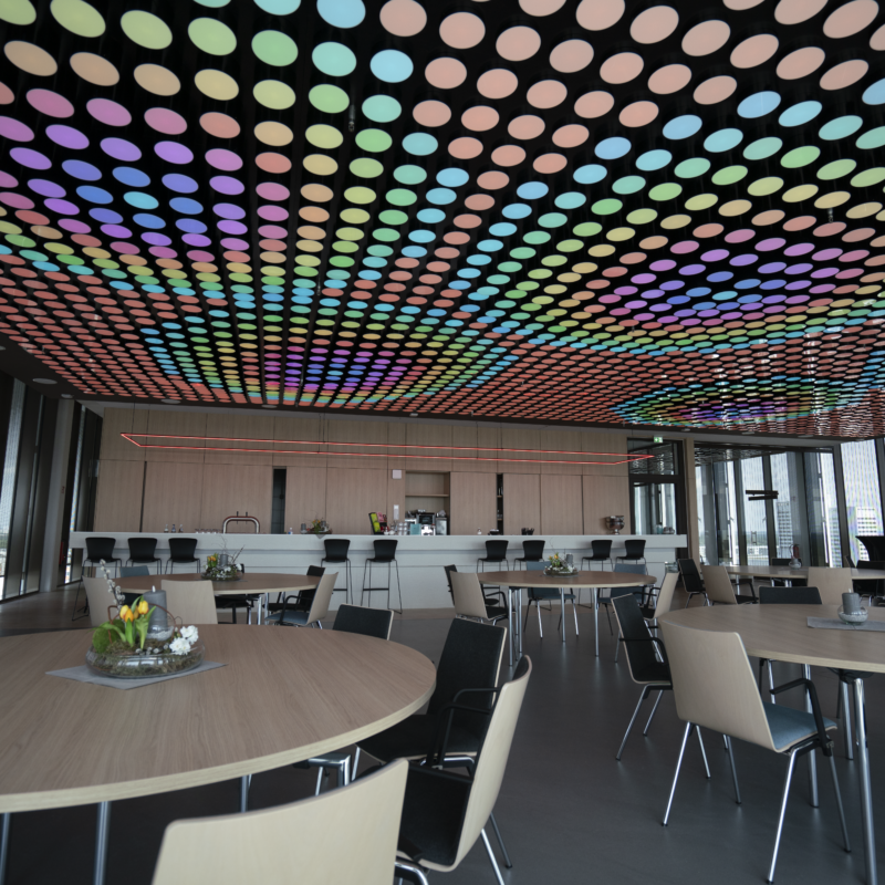 Ein moderner Raum mit einer auffälligen Lichtdecke, die aus zahlreichen farbigen, kreisförmigen Lichtern besteht. Die Decke leuchtet in verschiedenen Farbtönen, darunter Blau, Grün, Rosa und Gelb. Der Raum ist mit runden Holztischen und schwarzen sowie hellen Stühlen ausgestattet. Im Hintergrund befindet sich eine Theke, und große Fenster lassen viel Tageslicht herein.