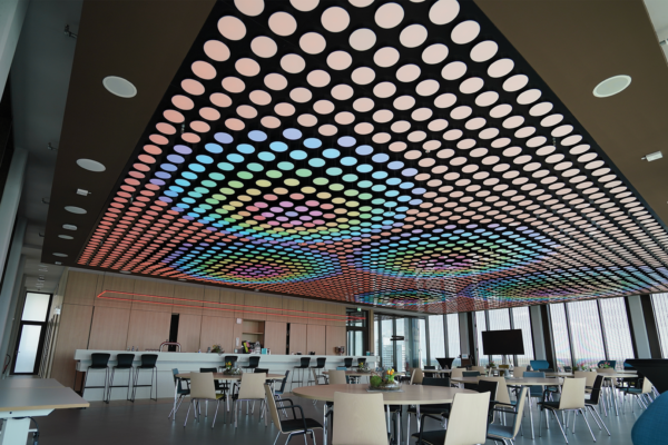 Moderner Raum mit einer auffälligen Lichtdecke, die aus bunten, kreisförmigen Lichtern besteht, die sich in einem Muster über die Decke erstrecken. Darunter befinden sich runde Holztische und moderne Stühle, während im Hintergrund eine Theke und große Fenster zu sehen sind, die Tageslicht in den Raum lassen.