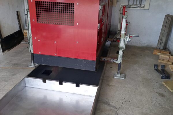 Ein roter Generator steht in einem technischen Raum an der Talsperre Sosa. Unter dem Generator befindet sich eine Auffangwanne aus Metall. An der Wand hängen Steuergeräte und Kabel. Der Raum ist funktional gestaltet, mit einem einfachen Betonboden und technischen Installationen.
