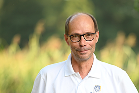 Portrait von Thomas Gruschka, einem Mitarbeiter von NEL Leipzig, in einem weißen Polohemd mit dem NEL-Logo. Er trägt eine Brille und steht vor einem unscharfen, grünen Hintergrund im Freien.