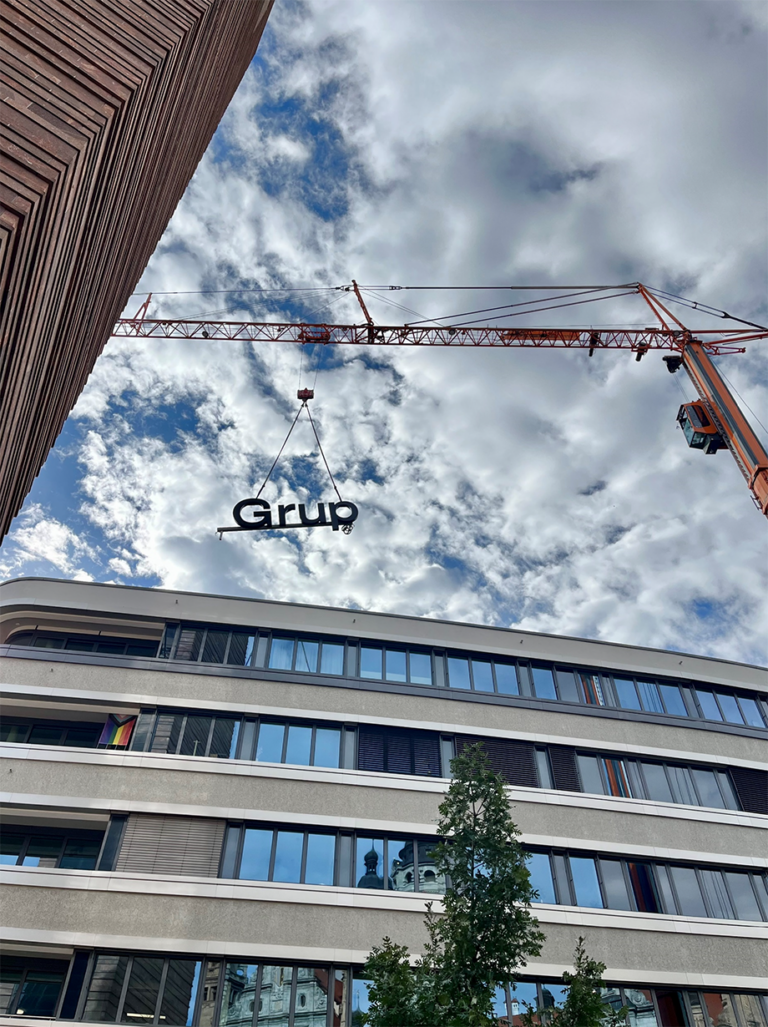 Ein großer Kran hebt den Schriftzug "Grup" über ein modernes Bürogebäude. Der Himmel ist leicht bewölkt, und das Gebäude im Vordergrund hat mehrere Fensterreihen. Eine Regenbogenfahne hängt an einem der Fenster.