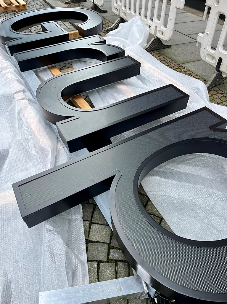 Große, schwarze 3D-Buchstaben der GRK Gruppe liegen auf dem Boden, offenbar zur Montage vorbereitet. Sie sind auf Folie und Holzplatten abgelegt, um sie vor Beschädigung zu schützen.