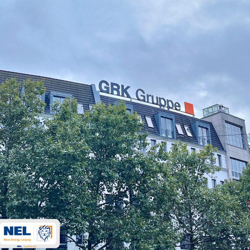 Außenansicht eines Bürogebäudes mit dem großen Schriftzug "GRK Gruppe" auf dem Dach. Ein Baum verdeckt teilweise die Fassade. Unten links ist das Logo von NEL (New Energy Leipzig) mit einem Löwenkopf sichtbar.