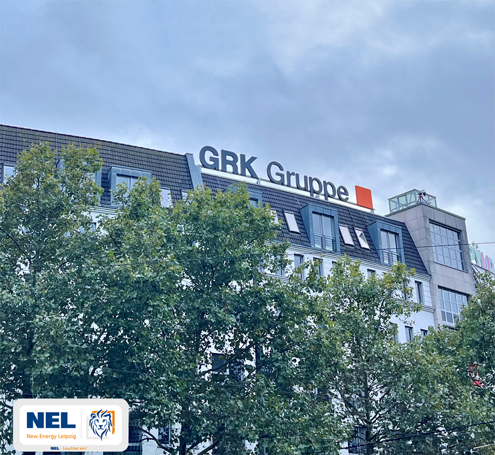 Außenansicht eines Bürogebäudes mit dem großen Schriftzug "GRK Gruppe" auf dem Dach. Ein Baum verdeckt teilweise die Fassade. Unten links ist das Logo von NEL (New Energy Leipzig) mit einem Löwenkopf sichtbar.