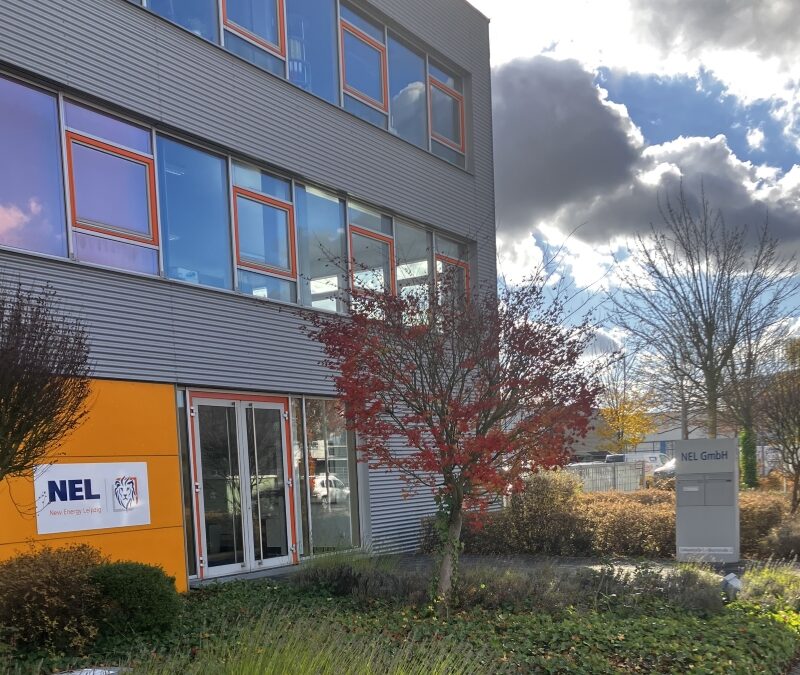 Außenansicht eines modernen Bürogebäudes von "NEL - New Energy Leipzig". Das Gebäude hat eine graue Fassade und mehrere Fensterreihen. Neben dem Eingang sieht man das Firmenlogo.. Vor dem Gebäude stehen einige Bäume und Sträucher, die den Eingang säumen. Im Vordergrund ist ein Weg sichtbar.