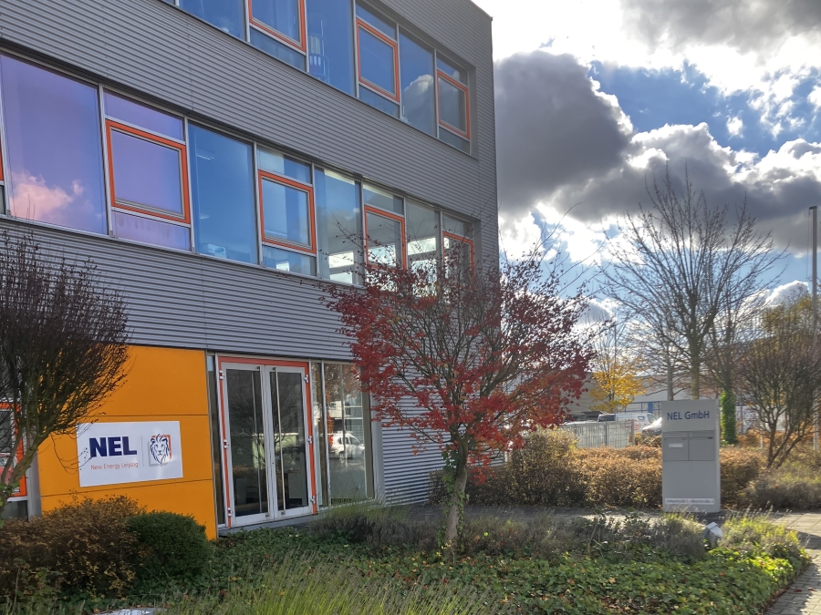 Außenansicht eines modernen Bürogebäudes von "NEL - New Energy Leipzig". Das Gebäude hat eine graue Fassade und mehrere Fensterreihen. Neben dem Eingang sieht man das Firmenlogo.. Vor dem Gebäude stehen einige Bäume und Sträucher, die den Eingang säumen. Im Vordergrund ist ein Weg sichtbar.
